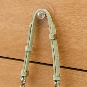 Puede incluir: Una correa de bolso ajustable de color verde claro con acabado texturizado, que presenta hebillas de metal dorado y un gancho de ventosa circular transparente con una pieza central de cristal facetado sobre una superficie de madera.