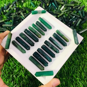 Op de afbeelding: Een verzameling van 17 groene edelsteenpunten, ook wel kristallen staven genoemd, gerangschikt op een wit oppervlak. De punten zijn in verschillende tinten groen, van donkergroen tot lichtgroen.