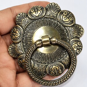 May include: A circular brass furniture pull featuring an intricate floral-inspired embossed pattern on the backplate and a textured, rope-style ring handle attached to the center. The metal has a polished, antique-toned finish with dark accents.