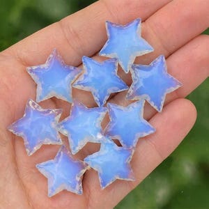 May include: A collection of nine opalescent star-shaped beads. Each star has a translucent, light blue center with a white, textured border. The beads are held in a hand, with a blurred green background.