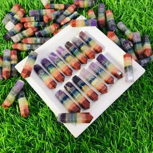 May include: A collection of seven chakra crystal wands, each with a different color representing a chakra. The wands are arranged on a white plate and are surrounded by green grass.
