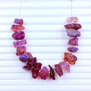 May include: A necklace featuring a strand of irregular, raw-cut gemstone chips in shades of deep purple, violet, and reddish-orange. The stones are separated by small, round, metallic silver-colored spacer beads and strung on a thin, clear wire.
