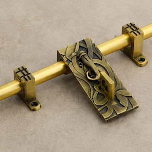 May include: A brass door bolt featuring a smooth cylindrical rod held by two square brackets with grid-patterned tops. A rectangular plate with an intricate, raised, dark-toned organic leaf-like pattern and a central latch mechanism is attached.