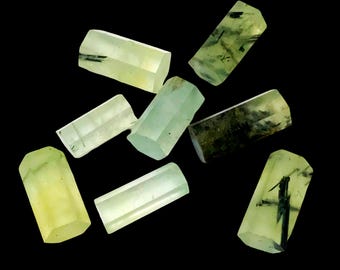 May include: A collection of translucent, light green prehnite crystals against a black background. The crystals vary in size and shape, with some displaying dark inclusions. The crystals have a faceted appearance.