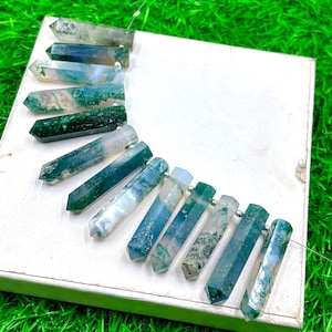 May include: A collection of polished moss agate crystal pendants. The pendants are a mix of green, blue, and white, with a hexagonal shape. They are strung together on a thin silver wire, and are displayed on a white surface.