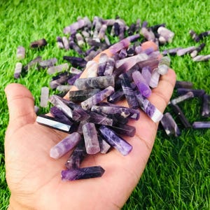 Può includere: Una mano che tiene un grande mucchio di punte di ametista. Le punte sono di varie tonalità di viola e alcune hanno un leggero luccichio.