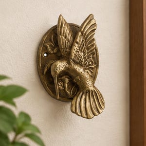 May include: A brass-toned metal door knocker featuring a three-dimensional hummingbird in flight. The bird is centered on an oval backplate decorated with embossed floral details. The piece has a weathered finish and a visible mounting hole.