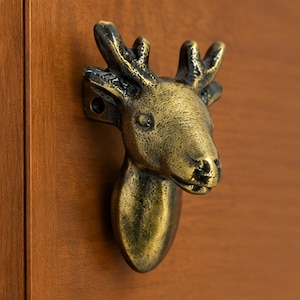 May include: A metallic cabinet knob shaped like a deer head with antlers, featuring a distressed antique brass finish with dark highlights. The hardware includes a visible mounting hole on the side, designed for attachment to a wooden surface.