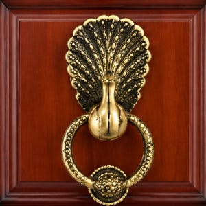 May include: A brass door knocker shaped like a peacock with an expansive, textured tail fan. The central body of the bird connects to a circular, beaded ring that hangs below, featuring a textured, spherical striking plate at the bottom center.