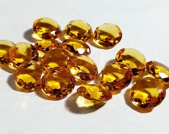 Puede incluir: Un conjunto de gemas facetadas de color amarillo dorado. Las gemas ovaladas presentan múltiples facetas, que capturan la luz y crean un efecto brillante. Las piedras están dispuestas sobre una superficie blanca, resaltando su color y claridad.