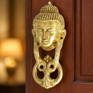 May include: A polished gold-colored metal door knocker shaped like a Buddha head with closed eyes and a textured crown. The knocker features a hinged, ornate circular ring mechanism attached below the face, mounted on a dark brown wooden door surface.