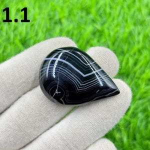 May include: A polished, teardrop-shaped agate gemstone in black and white. The stone displays a pattern of layered white and grey bands against a black base. The number "1.1" is visible in the top left corner.
