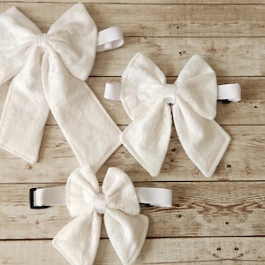 Może przedstawiać: Trzy białe, materiałowe muszki w różnych rozmiarach, z regulowanymi paskami. Muszki ułożone są na jasnobrązowej drewnianej powierzchni.