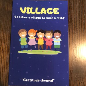 May include: A blue book with the title "VILLAGE" and the text "It takes a village to raise a child" in yellow. The book also has the text "Gratitude Journal" in yellow. The cover features a cartoon illustration of five children holding hands and smiling.