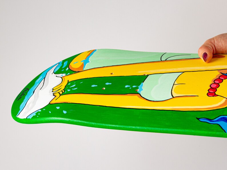 K&ouml;nnte beinhalten: Ein gr&uuml;nes Skateboard-Deck mit einem bemalten Design einer Person mit gelben Beinen und einem gr&uuml;n-wei&szlig;en Badeanzug. Die Person steht im Wasser mit einer wei&szlig;en Welle dahinter. Das Deck ist mit blauen und wei&szlig;en Wassertropfen bemalt.