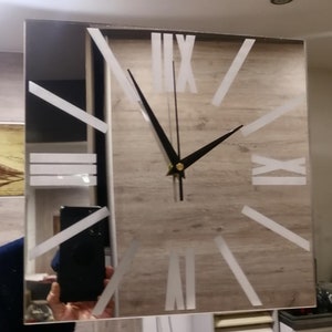 Op de afbeelding: Een spiegelklok met Romeinse cijfers en zwarte wijzers. De wijzerplaat van de klok is omgeven door een zilveren frame.