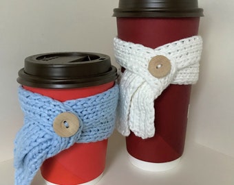 Puede incluir: Dos vasos de café desechables rojos con tapas marrones están envueltos en bufandas de punto. El vaso más pequeño tiene una bufanda azul claro con un botón de madera. El vaso más grande tiene una bufanda blanca con un botón de madera.