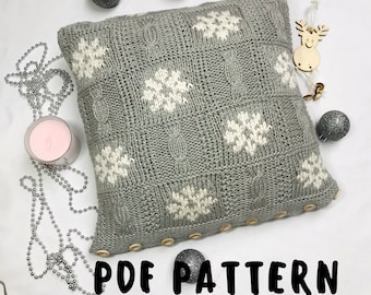 Puede incluir: Una almohada de punto gris con diseños de copos de nieve blancos y botones de madera. La almohada está sobre una superficie blanca con cuentas plateadas y una vela rosa. El texto "PDF PATTERN" está impreso debajo de la almohada.