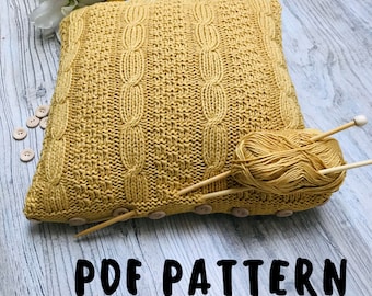 Puede incluir: Un patrón PDF para una almohada tejida con un diseño de punto de cable. La almohada es de color amarillo mostaza y se muestra con agujas de tejer de madera y una bola de hilo.