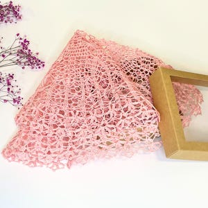 Puede incluir: Un encaje de ganchillo rosa con un delicado patr&oacute;n floral. El encaje est&aacute; doblado por la mitad y colocado sobre una superficie blanca. Una caja marr&oacute;n con una ventana transparente est&aacute; en el fondo.