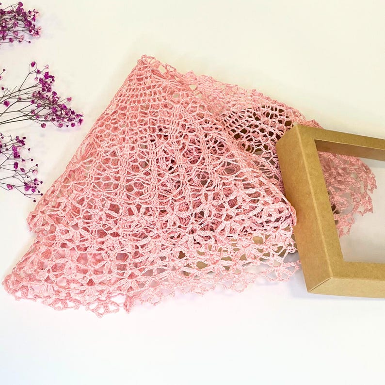 Puede incluir: Un encaje de ganchillo rosa con un delicado patr&oacute;n floral. El encaje est&aacute; doblado por la mitad y colocado sobre una superficie blanca. Una caja marr&oacute;n con una ventana transparente est&aacute; en el fondo.