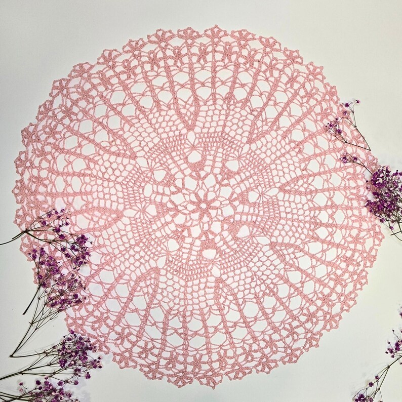Puede incluir: Un delicado encaje de ganchillo rosa con un dise&ntilde;o circular. El encaje tiene un patr&oacute;n intrincado con un motivo floral central.