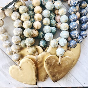 May include: A garland of blue, green, and beige wooden beads with gold accents hangs with three gold heart-shaped ornaments.