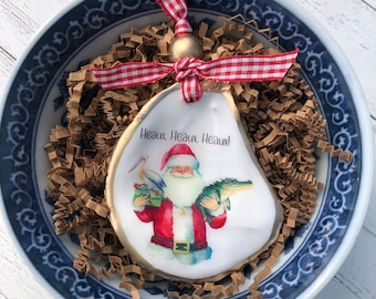 May include: A white oyster shell Christmas ornament with a gold rim. The ornament features a watercolor illustration of Santa Claus holding a gift and an alligator. The text "Heaux, Heaux, Heaux!" is printed on the ornament.