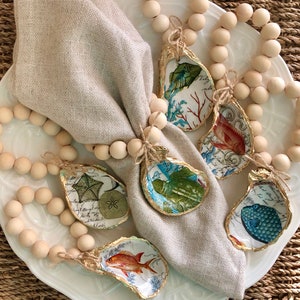 May include: A set of six oyster shell napkin rings with a variety of nautical designs. The rings are strung on a wooden bead garland.