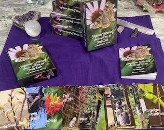 May include: An oracle deck with the title "Empower Yourself" featuring a butterfly on a flower. The deck includes a guidebook and individual cards with nature-themed images and words like "Conditional Love" and "Strong Truth". The items are arranged on a purple cloth.