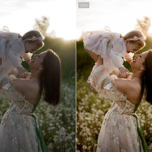 May include: A woman holding a young child in a field of white flowers. The woman wears a light-colored dress with floral embroidery and a green sash. The child is wearing a white dress with a pink bow. The image is bathed in warm sunlight.