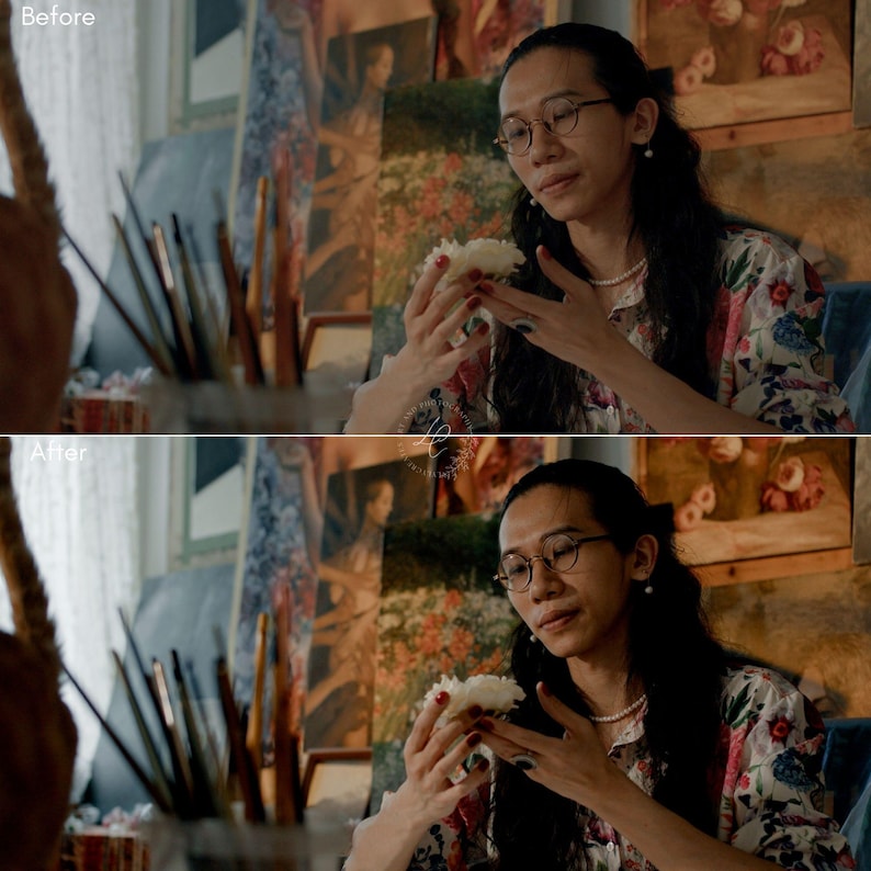 May include: A person wearing glasses and a floral shirt holds a white flower. The background includes paintings and art supplies. The person is wearing a pearl necklace and has long, dark hair.
