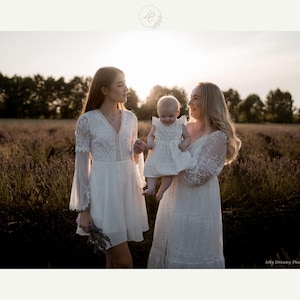 May include: Three people in white dresses stand in a field of lavender at sunset. One woman holds a baby in a white dress. The dresses have lace and flowing sleeves. The scene is bathed in warm sunlight, creating a soft, romantic atmosphere.