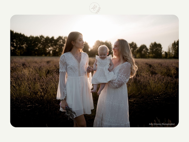 May include: Three people in white dresses stand in a field of lavender at sunset. One woman holds a baby in a white dress. The dresses have lace and flowing sleeves. The scene is bathed in warm sunlight, creating a soft, romantic atmosphere.