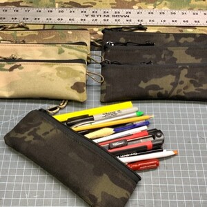 May include: Three zippered pouches in camouflage patterns, two in a green and brown pattern and one in a black and green pattern. The pouches are filled with writing utensils, including pencils, pens, and a utility knife.