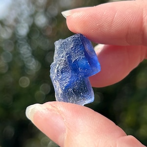 Puede incluir: Un pequeño cristal azul oscuro, tallado en bruto, se sostiene entre los dedos sobre un fondo exterior borroso. La forma irregular y el color vibrante del cristal son el foco. Se ve un anillo plateado con un diseño celestial.