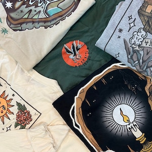 May include: Five different t-shirts with various graphic designs. The designs include a pair of hiking boots, a bird with a flower, a mountain scene, a sun with flowers, and a candle in a cave.