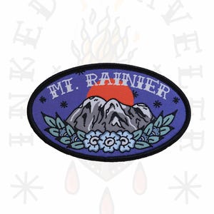 Puede incluir: Un parche bordado de color morado y azul con una cordillera montañosa, un sol rojo y estrellas blancas. El texto "MT. RAINIER" está escrito en blanco en el parche.
