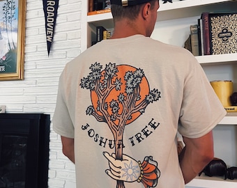 May include: A beige t-shirt with a graphic of a Joshua Tree in a hand with a sun and a crown. The text "Joshua Tree" is printed below the image.