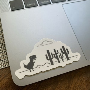 May include: A black and white sticker of the classic dinosaur game from Google Chrome. The dinosaur is running across a pixelated desert landscape with a cloud above and cacti in the background.