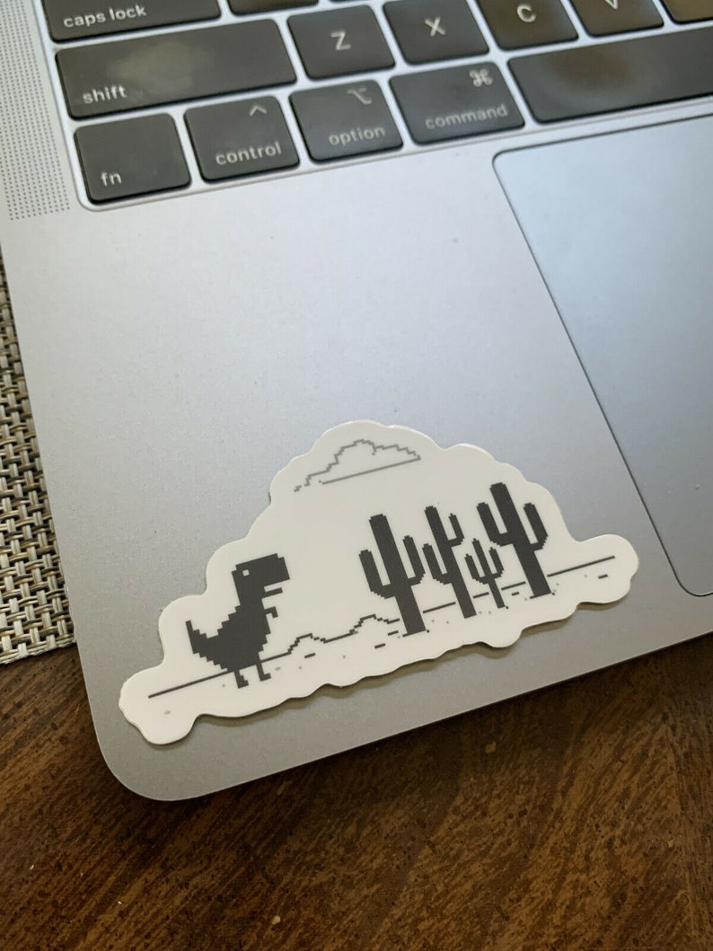May include: A black and white sticker of the classic dinosaur game from Google Chrome. The dinosaur is running across a pixelated desert landscape with a cloud above and cacti in the background.