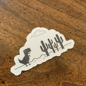 May include: A white sticker with a black and white pixelated image of a dinosaur running through a desert landscape with a cloud in the sky. The dinosaur is running towards a row of cacti.