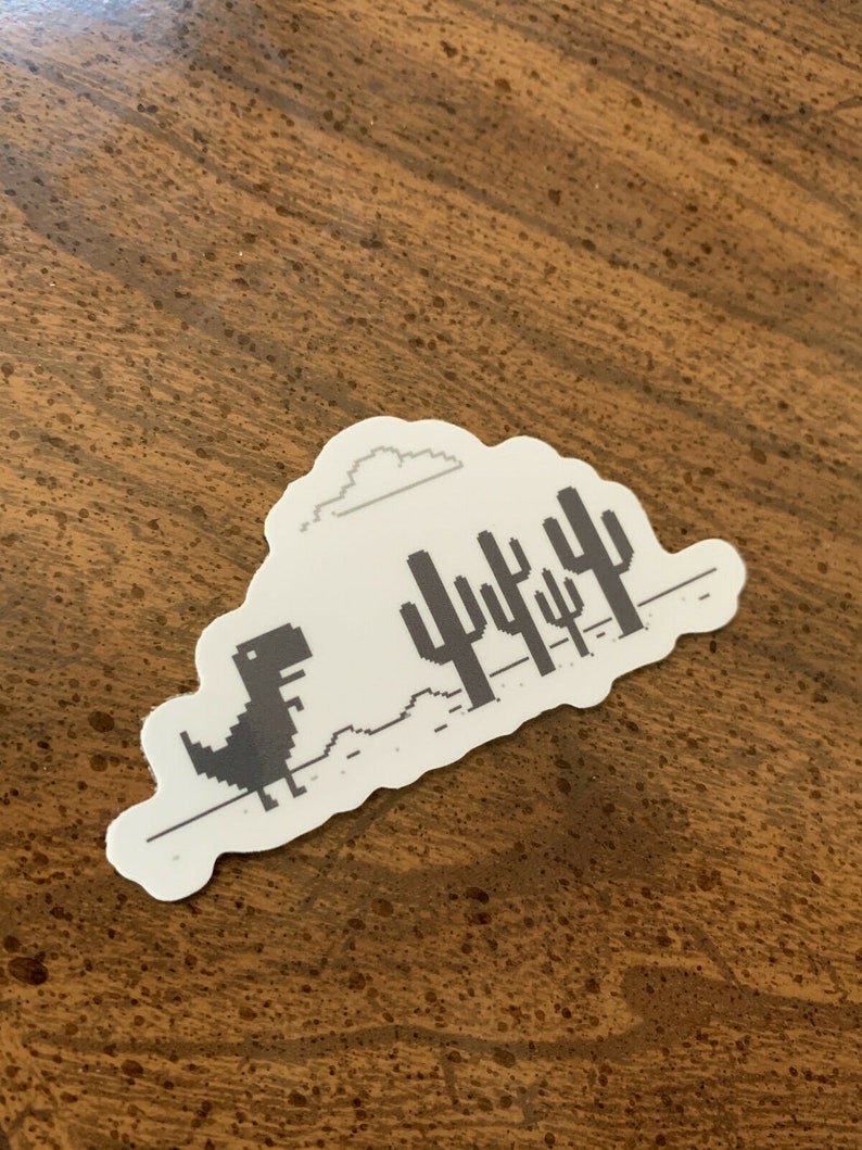 May include: A white sticker with a black and white pixelated image of a dinosaur running through a desert landscape with a cloud in the sky. The dinosaur is running towards a row of cacti.