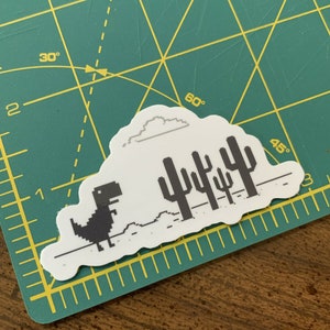 May include: A white sticker with a black pixelated dinosaur running across a white landscape with a gray cloud and black pixelated cacti. The sticker is on a green cutting mat.