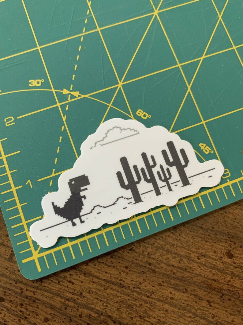 May include: A white sticker with a black pixelated dinosaur running across a white landscape with a gray cloud and black pixelated cacti. The sticker is on a green cutting mat.