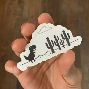 May include: A white sticker with a black and white pixelated image of a dinosaur running through a desert landscape with cacti. The dinosaur is running to the right and there is a cloud above it.