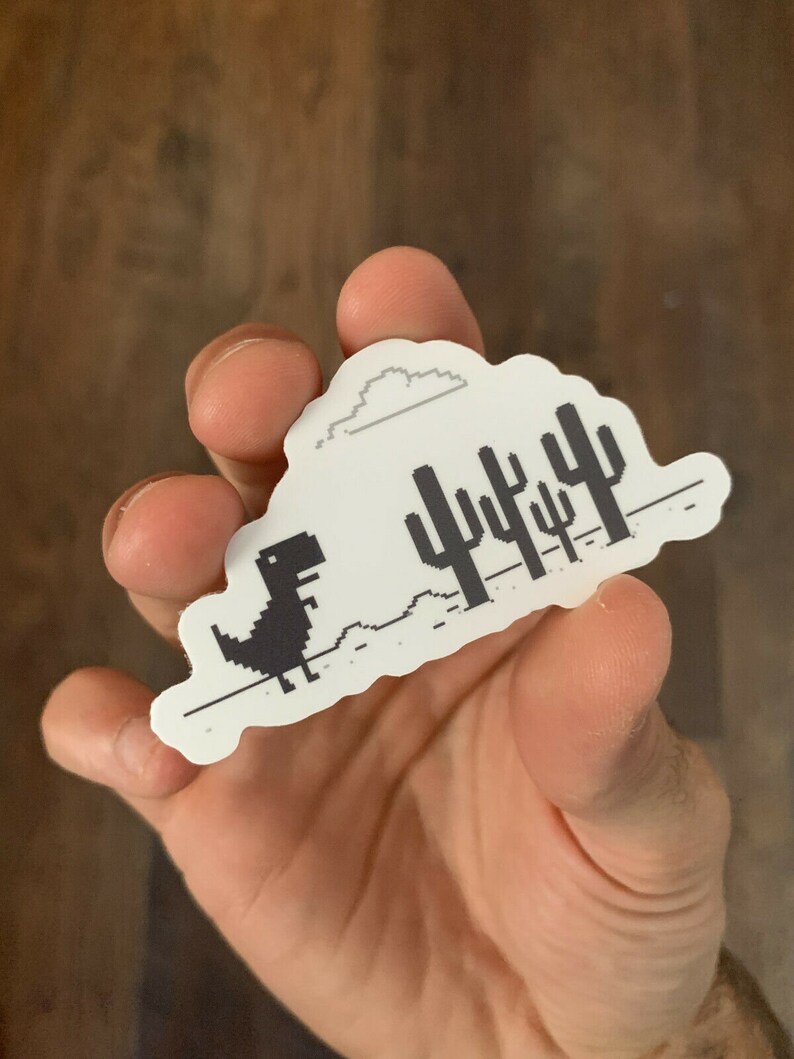 May include: A white sticker with a black and white pixelated image of a dinosaur running through a desert landscape with cacti. The dinosaur is running to the right and there is a cloud above it.