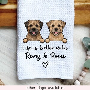 May include: A white waffle-textured kitchen towel featuring a graphic of two brown Border Terrier dogs with their paws resting on a line. Below the dogs, the black text reads Life is better with Remy & Rosie, followed by a small black heart symbol.