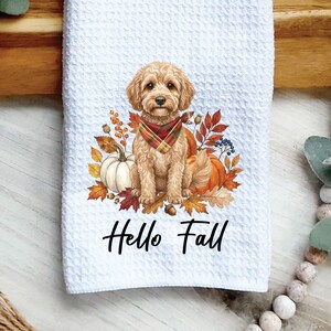May include: A white waffle-textured kitchen towel featuring a central illustration of a golden-colored doodle dog wearing a red, green, and yellow plaid bandana, surrounded by autumn leaves, pumpkins, and berries, with the text Hello Fall below.