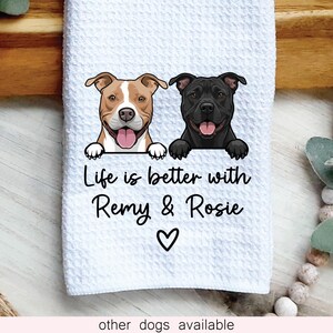 May include: A white waffle-textured kitchen towel featuring a graphic of a tan and white dog and a black dog with their paws resting on a ledge. Below the dogs, the text reads Life is better with Remy & Rosie, followed by a small black heart.